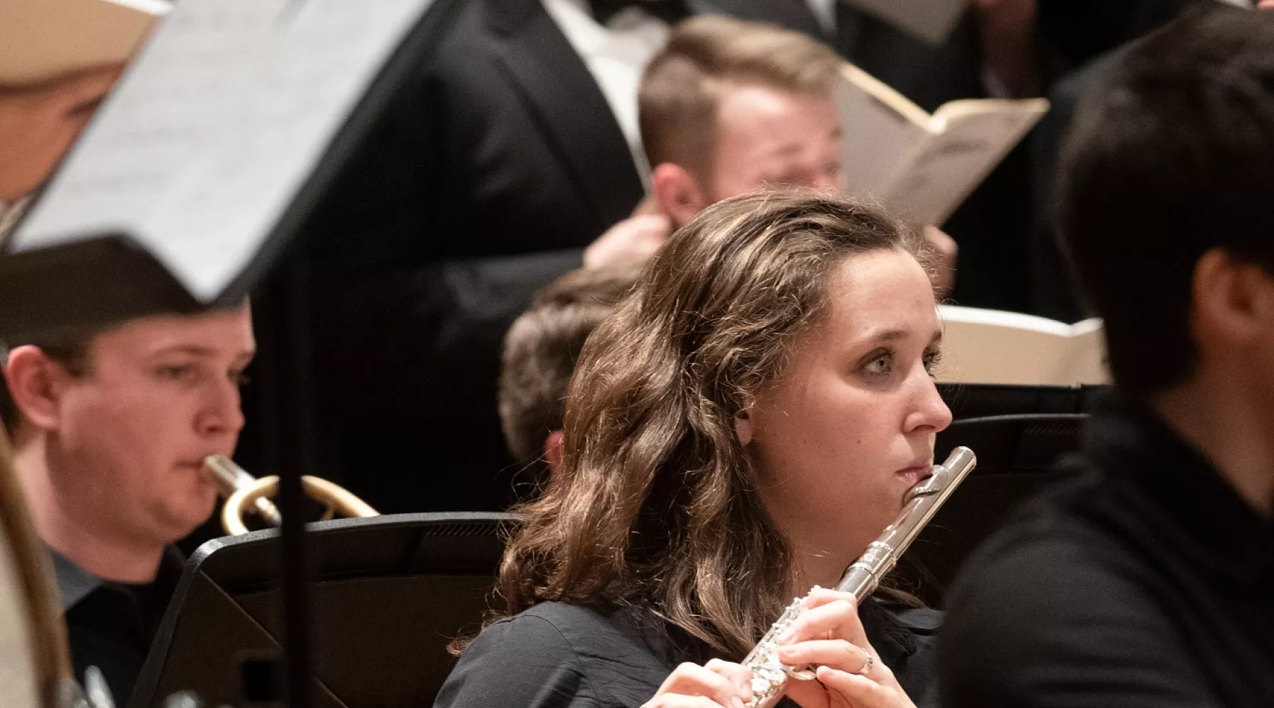 Student playing the flute with singers in the background at a music symphony concert.